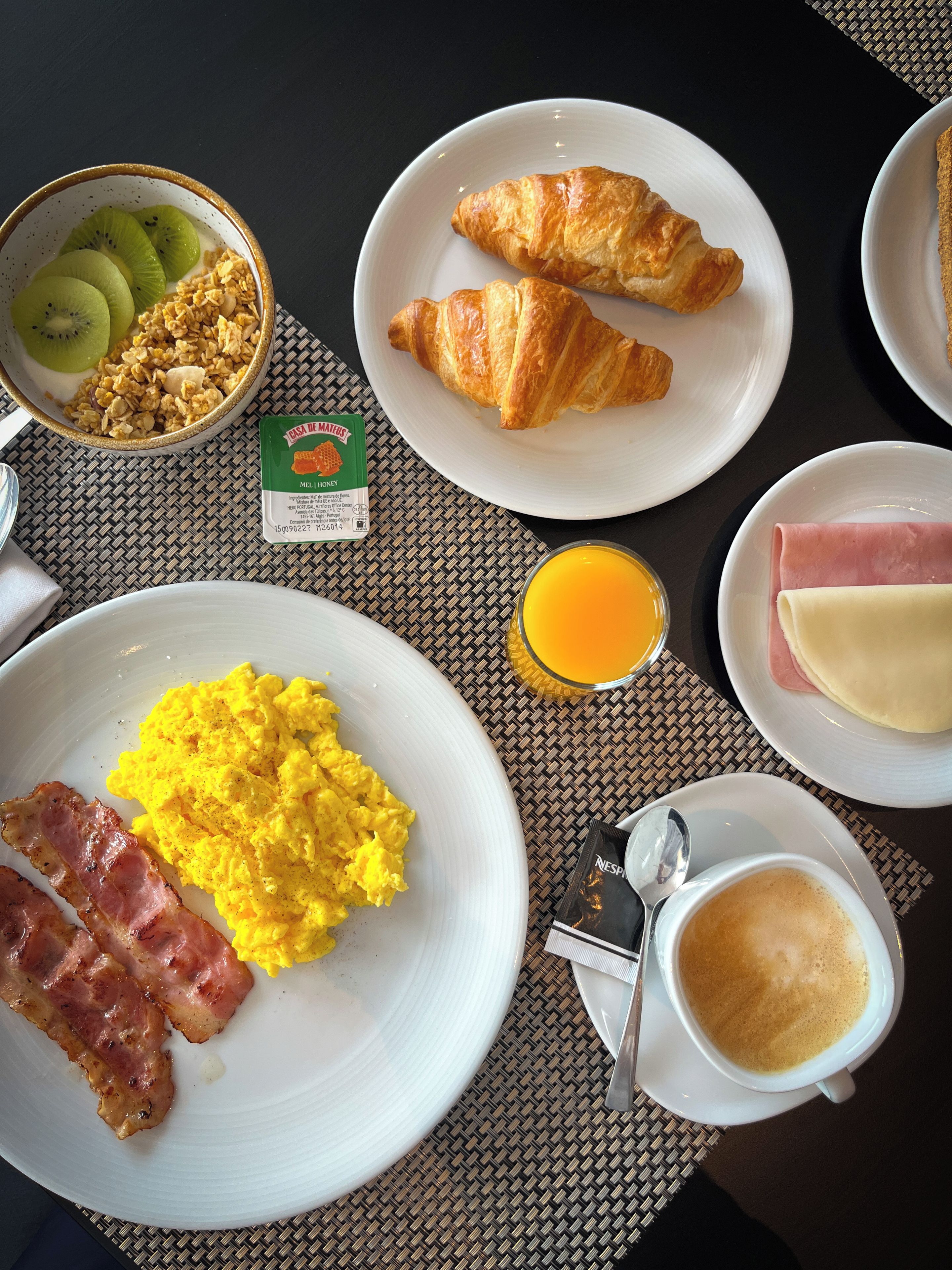 Una mesa de desayuno con huevos revueltos, tocino, croissants y un tazón de cereal con kiwi.