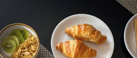 Una mesa de desayuno con huevos revueltos, tocino, croissants y un tazón de cereal con kiwi.