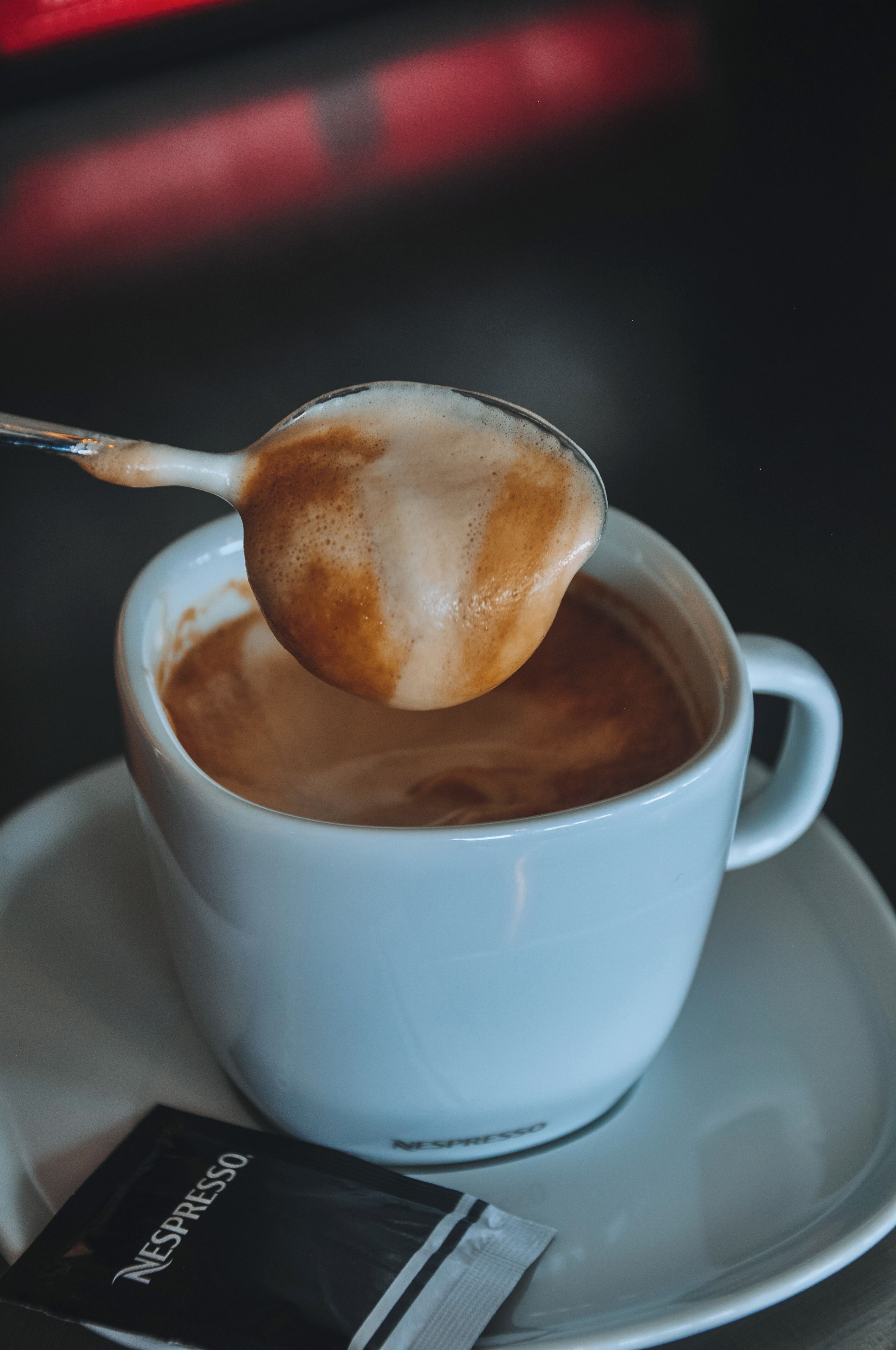 Una taza blanca de café con una cuchara levantando espuma, un platito y un paquete de cápsulas Nespresso.