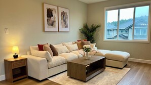 Un salon moderne avec un canapé beige, une table basse en bois et une grande fenêtre donnant sur des maisons.