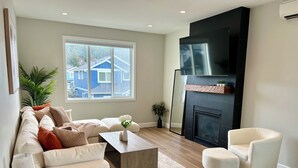 Un salon moderne comprenant un canapé, une table basse et un fauteuil, une cheminée et une grande fenêtre donnant sur des maisons.