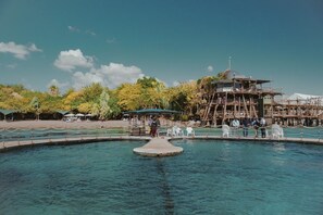 Ein klares Schwimmbecken mit einer zentralen Insel, umgeben von einer Holzterrasse und einer Aussichtsturm-artigen Struktur.