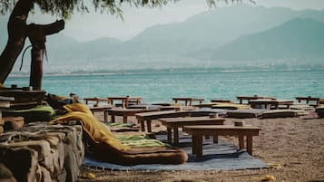 Ein Strand mit Holztischen und -bänken, einem Baum und Blick auf Meer und Berge im Hintergrund.