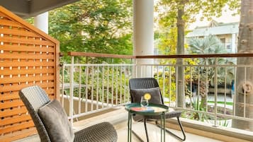 Ein Balkon mit zwei Korbstühlen, einem Glastisch mit einem Buch und einem Glas sowie Blick auf einen Garten und Gebäude.