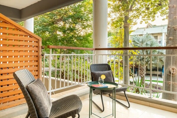 A balcony with two wicker chairs, a glass table with a book and a glass, and a view of a garden and buildings.