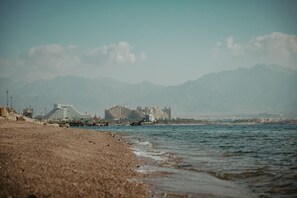 A beach with buildings in the background, a clear sky, and mountains in the distance.