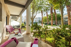 Un área de estar al aire libre techada, con sillas y una mesa, rodeada de palmeras y vistas a la calle.