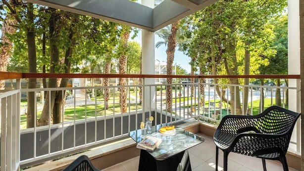 A balcony with a table and chairs, overlooking a green landscape with trees and a road. - Bright 1BR Royal Park Apt with Garden View (Eilat)