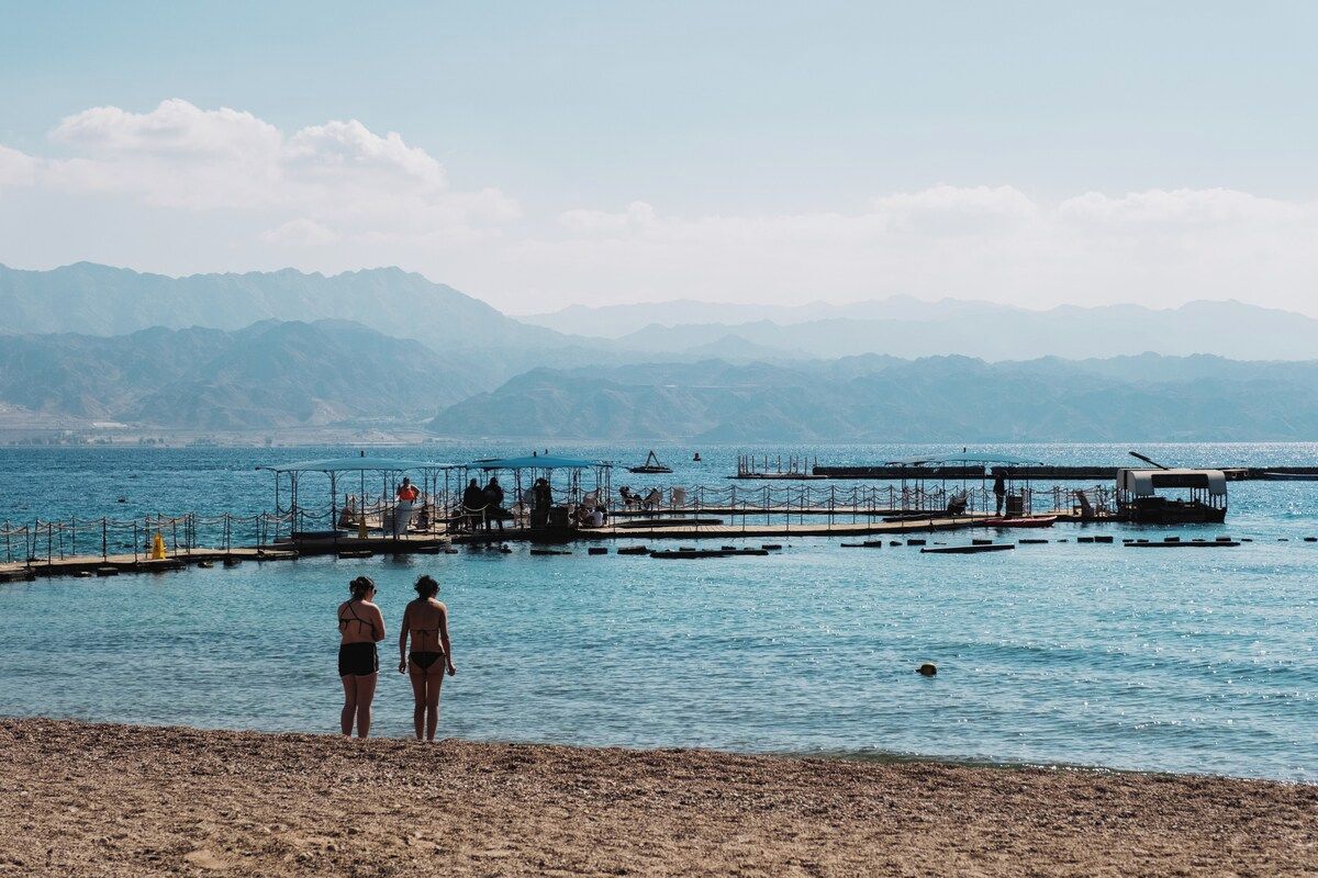 A beach with two people standing on the shore, a pier extending into the water, and mountains in the background.