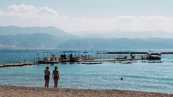 A beach with two people standing on the shore, a pier extending into the water, and mountains in the background.
