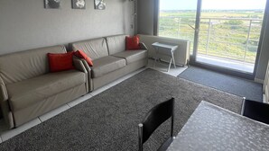 A modern living room with a grey sofa, red cushions, a small white table, and a large window offering a view of the countryside. - Studio in Middelkerke With Pool and Sea View (Middelkerke)