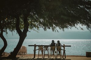 Terraza de madera con sillas dando vista a un cuerpo de agua, un árbol proporcionando sombra y una vista distante de montañas.