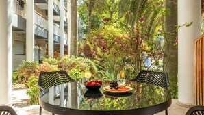 Una mesa con cubierta de vidrio y dos sillas, un cuenco de fruta y un vaso de agua en un entorno exterior con vistas a un edificio y exuberante vegetación.