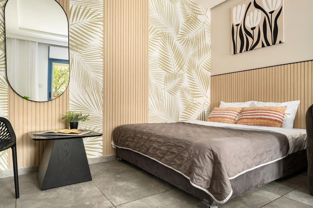 A bedroom with a bed, a bedside table with a book and a plant, a mirror, and a wall with a leaf pattern.