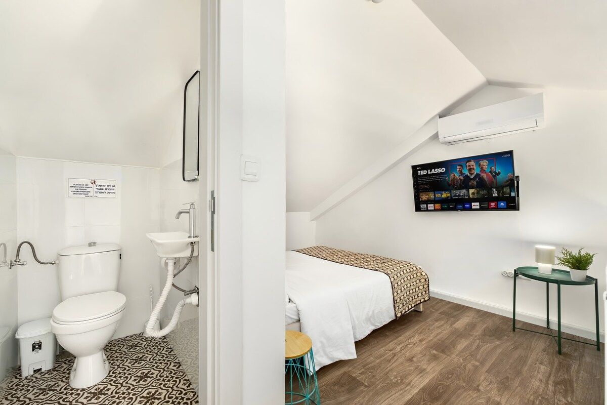 Ein modernes Badezimmer mit Toilette, Waschbecken und einem gemusterten Boden. Nebenan befindet sich ein Schlafzimmer mit einem Bett, einem Nachttisch und einem Fernseher, der einen Film zeigt.