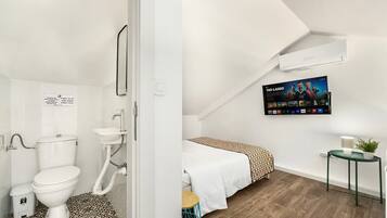 Ein modernes Badezimmer mit Toilette, Waschbecken und einem gemusterten Boden. Nebenan befindet sich ein Schlafzimmer mit einem Bett, einem Nachttisch und einem Fernseher, der einen Film zeigt.