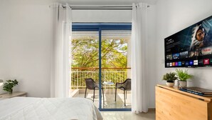Habitación de hotel con cama, televisión montada en la pared, cómoda de madera y balcón con vista a árboles.