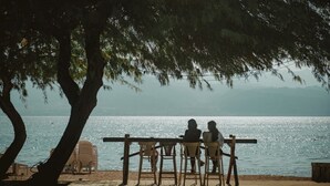 Une terrasse en bois avec des chaises donnant sur une étendue d’eau, un arbre offrant de l’ombre et une vue lointaine sur des montagnes.