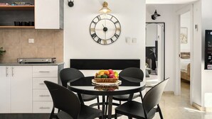 A modern kitchen with white cabinetry, a round dining table with black chairs, and a wall-mounted clock. - Bright 2BR Apartment with Balcony at Royal Park (Eilat)