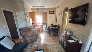 Una sala de estar con sofá, mesa de centro y televisor. Se observa una cocina al fondo, con armarios de madera y una zona de comedor.