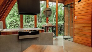 A modern wooden cabin with a stone fireplace, a glass door leading to a balcony, and a large window overlooking a forest. - CHALÉS SUIÇOS - Românticos na Natureza para Adultos (Itapecerica da Serra)