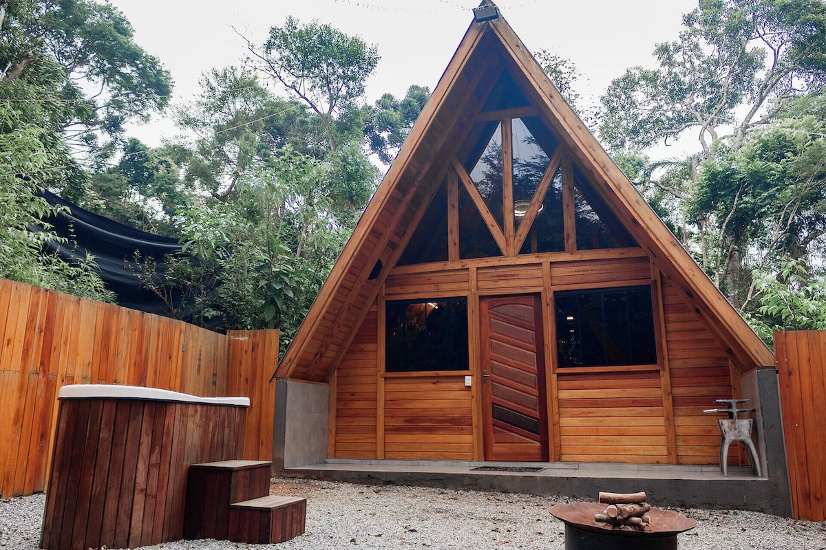 A wooden A-frame cabin with a large sliding door, a small porch, and a fire pit in front.