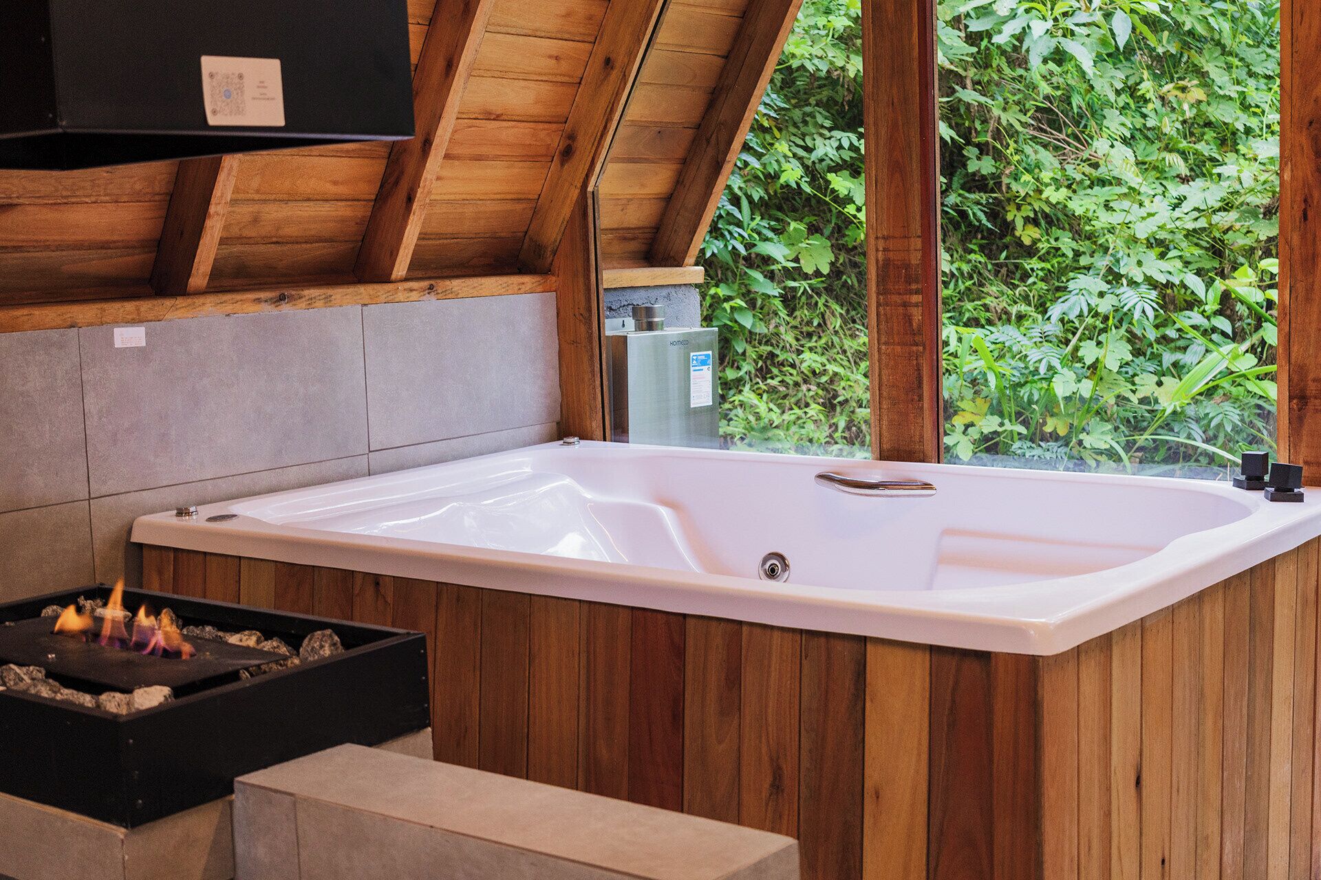 Un área de spa con una tina blanca grande, paneles de madera, un hogar y vistas a la vegetación a través de una ventana.