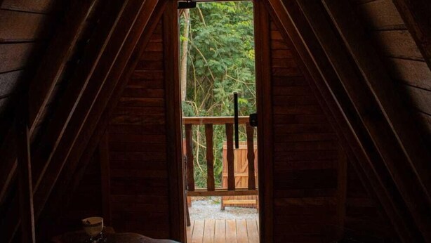An attic room with a sloped ceiling, a wooden table, and a window leading to an outdoor deck. - CHALÉS SUIÇOS - Românticos na Natureza para Adultos (Itapecerica da Serra)
