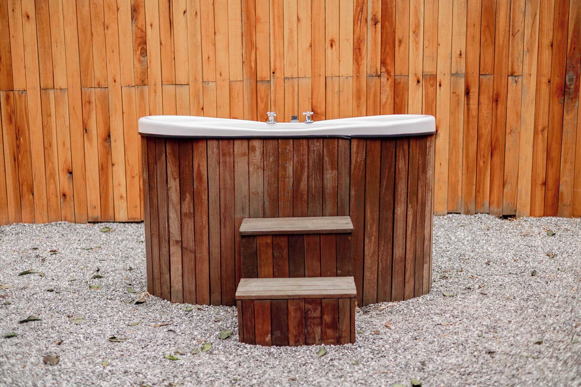 A wooden hot tub with a white interior and two steps leading up to it, set against a wooden fence background.