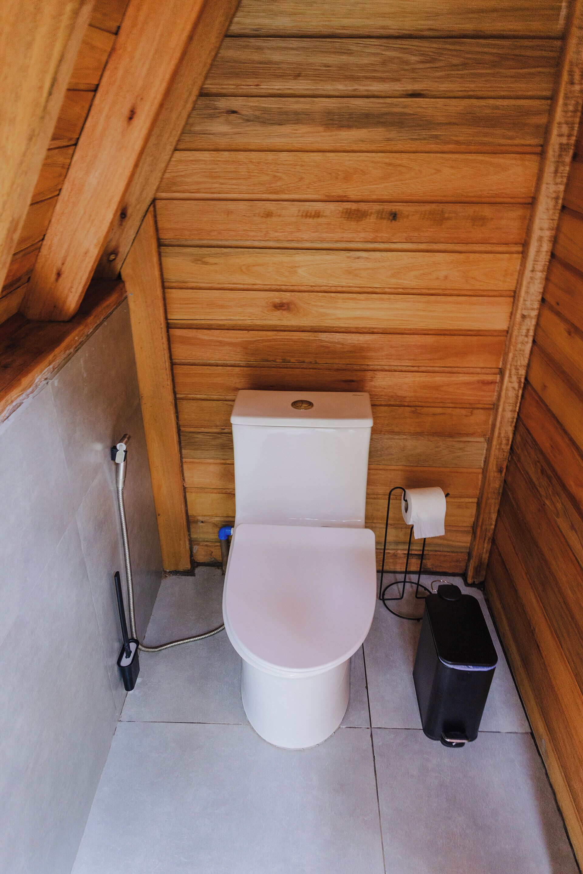 A small, wooden-paneled bathroom with a white toilet, a bidet, and a black trash bin.