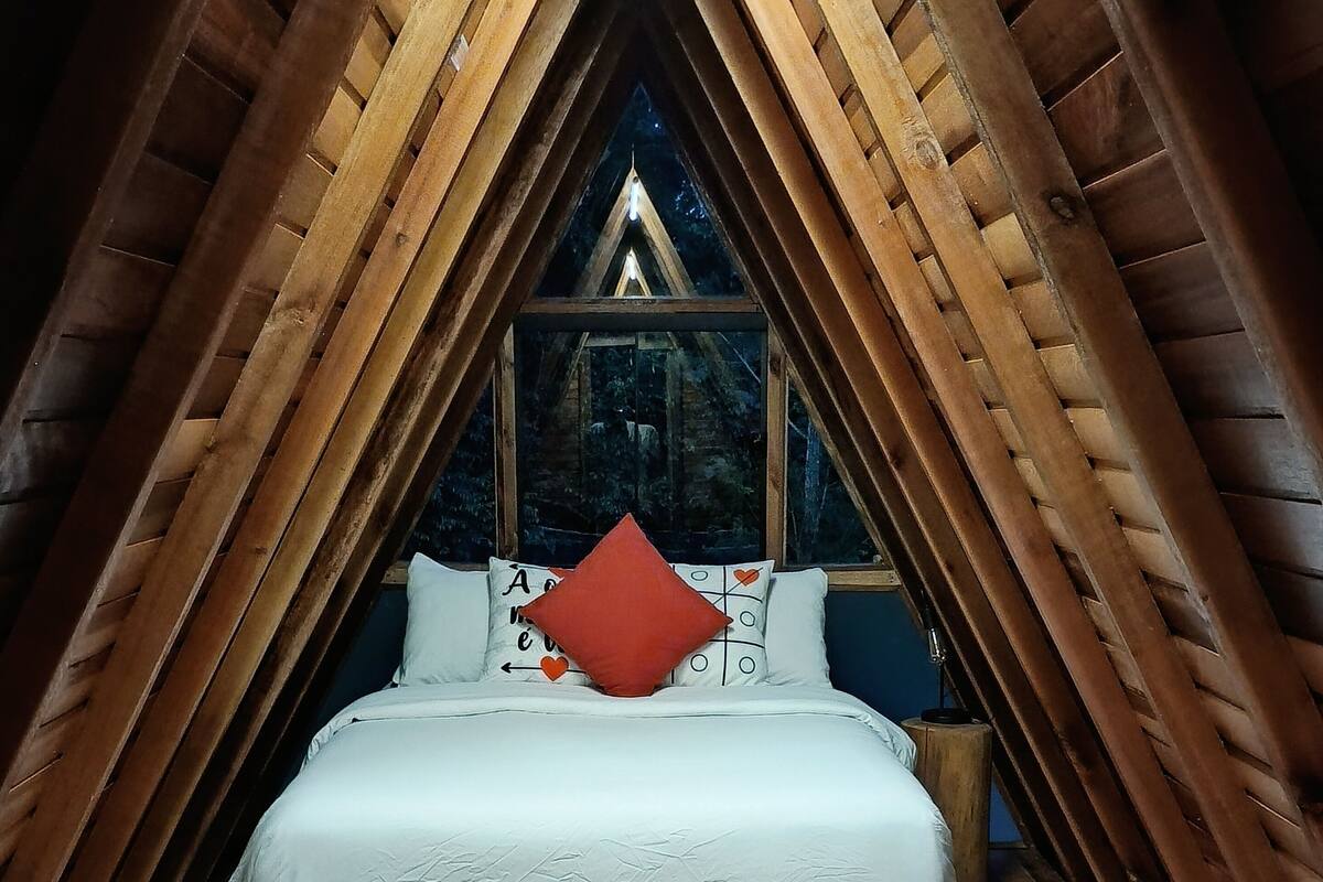 A cozy, triangular wooden room with a bed centered under a window, wooden slats on the ceiling, and a view of trees outside.