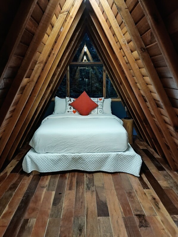 A cozy, triangular wooden room with a bed centered under a window, wooden slats on the ceiling, and a view of trees outside. - CHALÉS SUIÇOS - Românticos na Natureza para Adultos (Itapecerica da Serra)