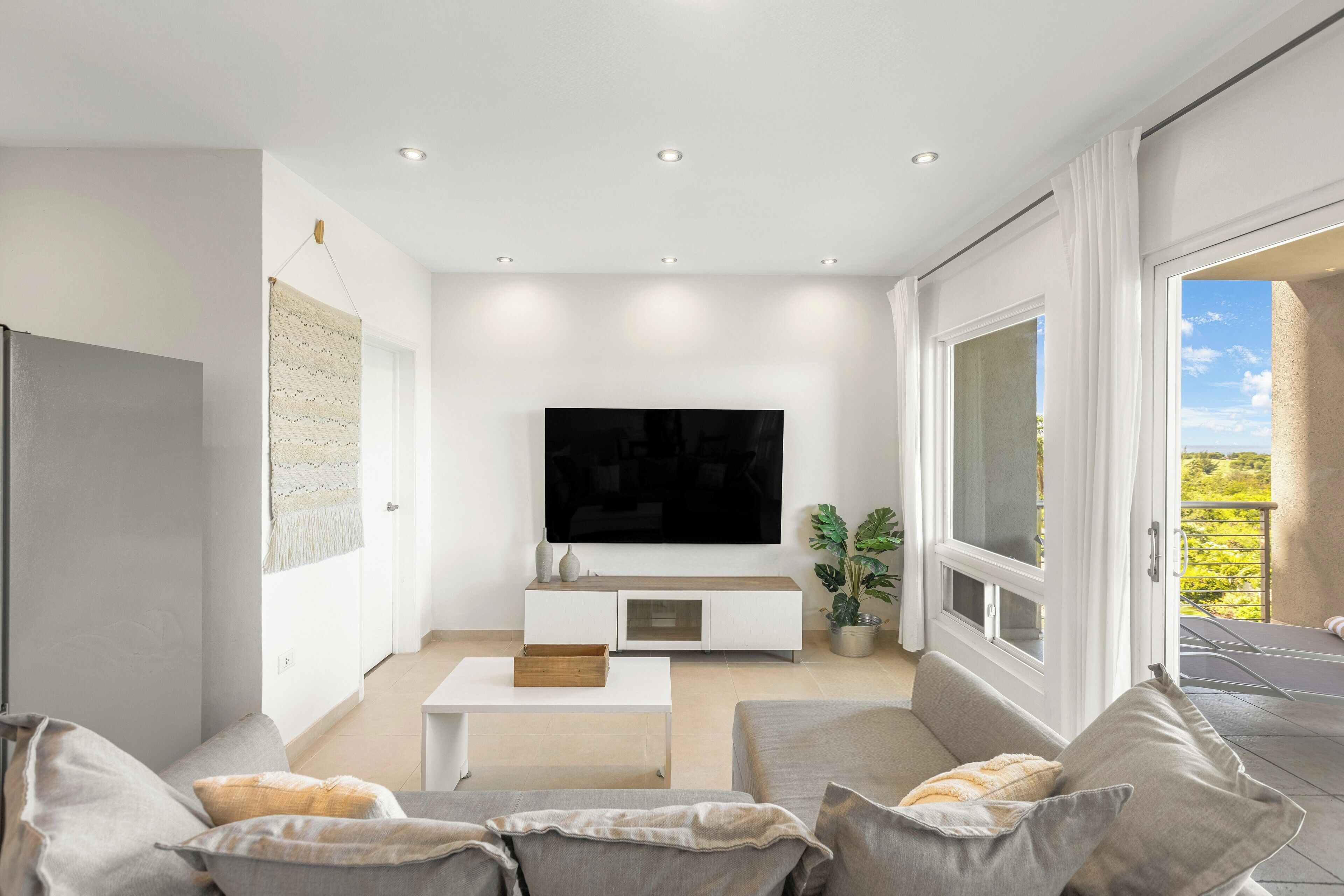 A modern living room with a flat-screen TV, a white entertainment unit, and a large window offering a view of the outdoors.