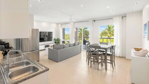 A modern kitchen with a stainless steel refrigerator, a built-in oven, and a sink. There is a grey sofa and a dining table with chairs in the living area. - Azure Heaven, Breathtaking views, Pool, Gym (Maho)