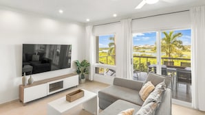 A modern living room with a flat-screen TV, a sofa, a coffee table, and large windows offering a view of a landscaped area. - Azure Heaven, Breathtaking views, Pool, Gym (Maho)