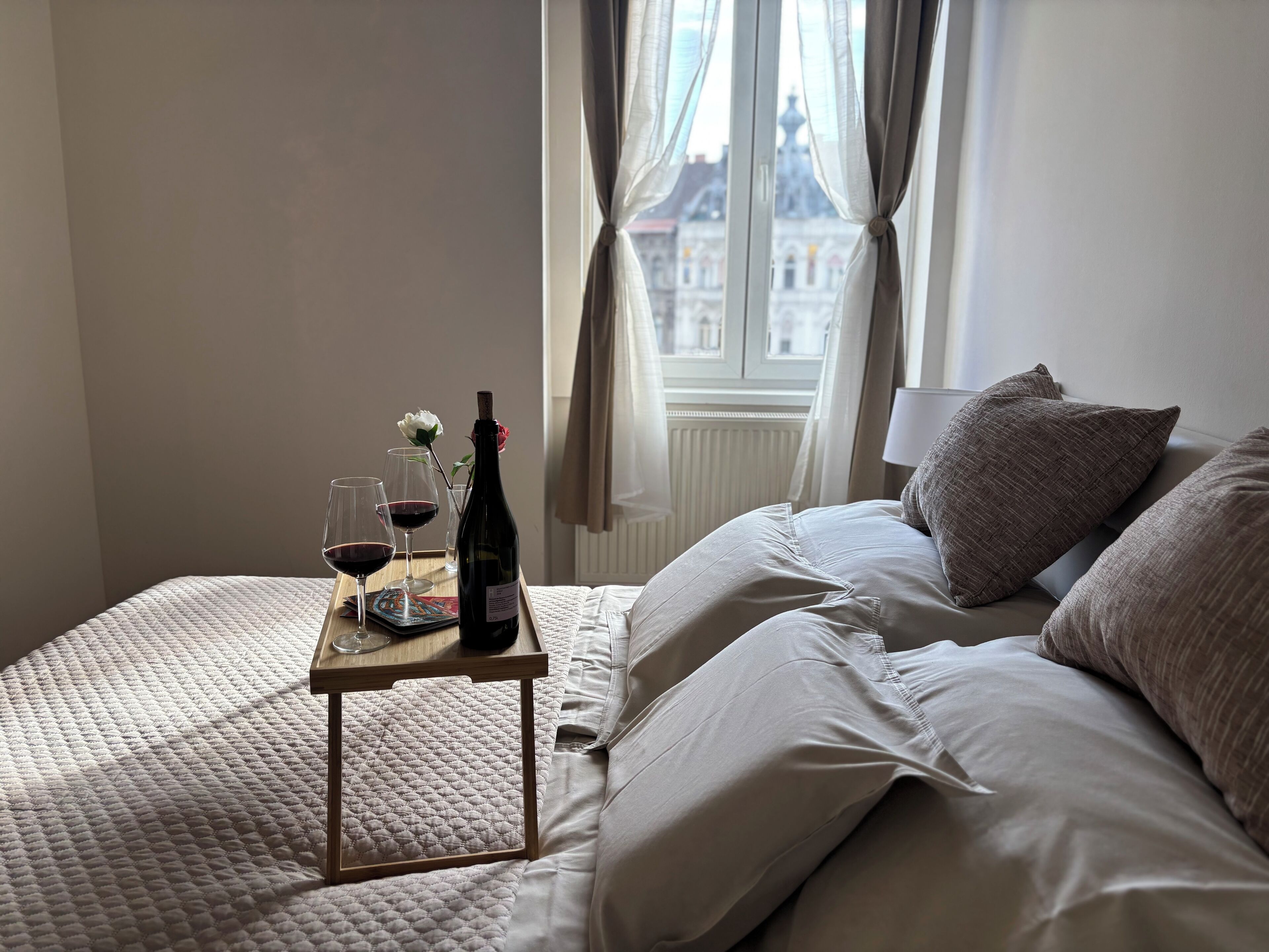 A neatly made bed with a small table beside it, holding a wine bottle, two wine glasses, and a single white flower.