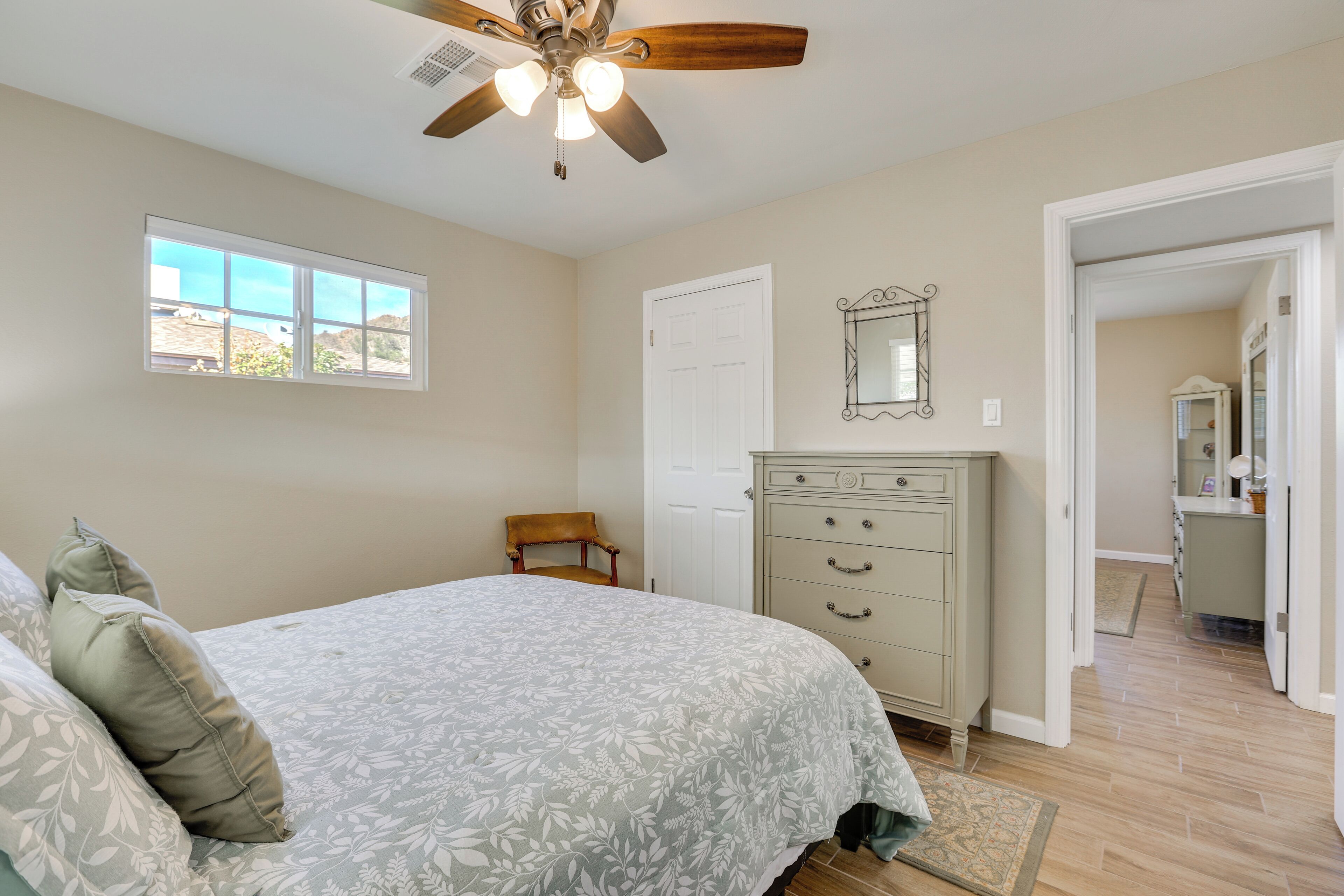 Une chambre à coucher comprenant un lit, une commode, un ventilateur de plafond, une fenêtre et une porte donnant sur une autre pièce.