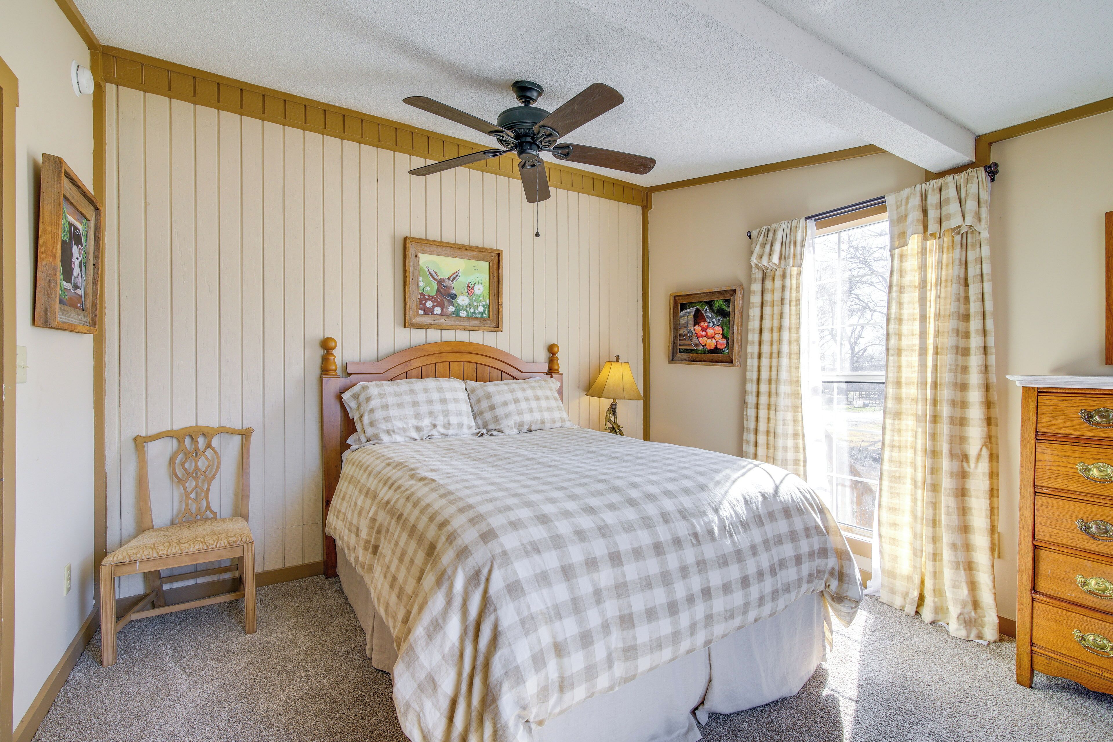A bedroom with a bed, a wooden chair, a ceiling fan, a dresser, and a window with curtains.