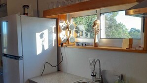 A compact kitchen with a refrigerator, sink, and stove. There is a window above the sink, and a shelf with glassware.