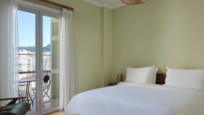 A hotel room with a large bed, a chair, a small table, and a view of the city through a sliding glass door.