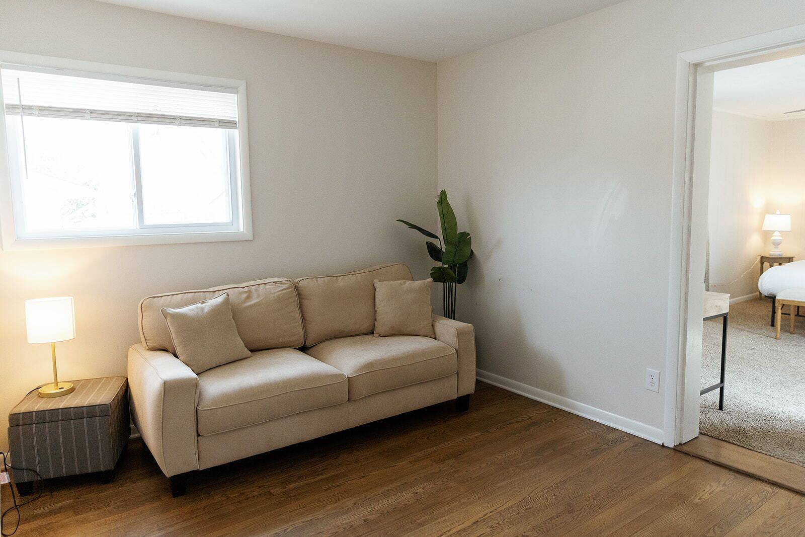 Ein Wohnzimmer mit einem beigen Sofa, einem kleinen Beistelltisch mit Lampe, einem Blumentopf und Holzboden.