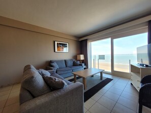 Ein modernes Hotelzimmer mit grauem Sofa, einem kleinen Tisch und einem Fernseher. Ein großes Fenster bietet Blick auf den Strand.