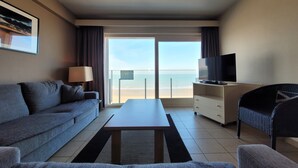 Ett hotellrum med en soffa, en tv på ett stativ, ett soffbord och utsikt över stranden.