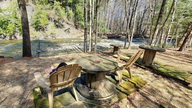 Un espace extérieur aménagé avec une table ronde en bois et des chaises, entouré d’une zone boisée, avec une rivière en arrière-plan.