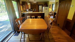 Une table à manger en bois avec six chaises, un coin cuisine avec des meubles en bois et une grande fenêtre avec des rideaux.