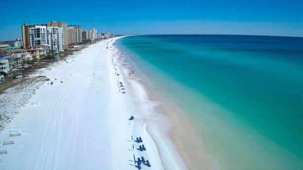 An aerial view of a beach with a long stretch of white sand, a clear blue ocean, and a row of buildings along the shore.