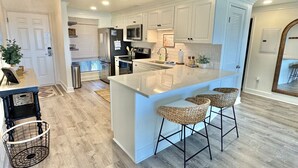 A modern kitchen with white cabinetry, a large island with bar stools, stainless steel appliances, and a chandelier.