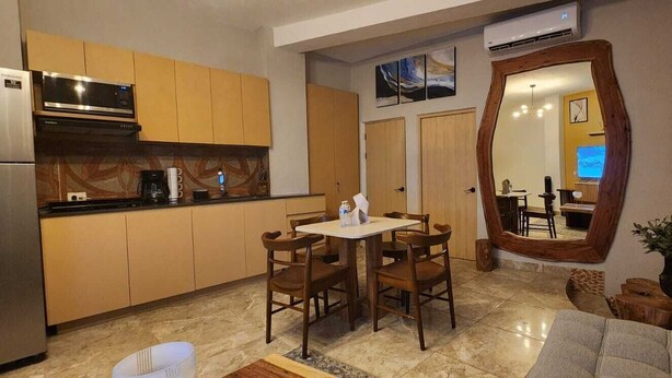 A compact kitchen with wooden cabinets, a dining table with chairs, a large mirror, and a living area with a sofa and a plant. - Apartment in downtown San Jose del Cabo on the transpeninsular highway. (San José del Cabo)