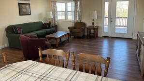 Un salon avec un canapé vert, une chaise en bois, une table en bois, un lit avec une tête de lit en bois, un ventilateur de plafond et une porte donnant sur l’extérieur.