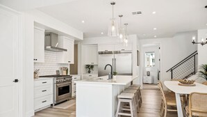 A modern kitchen with white cabinetry, a central island, stainless steel appliances, and a dining area with wicker chairs. - Sunstone Retreat: Hot Tub + Desert Color Lagoon + Beaches, Hot Tub (St. George)